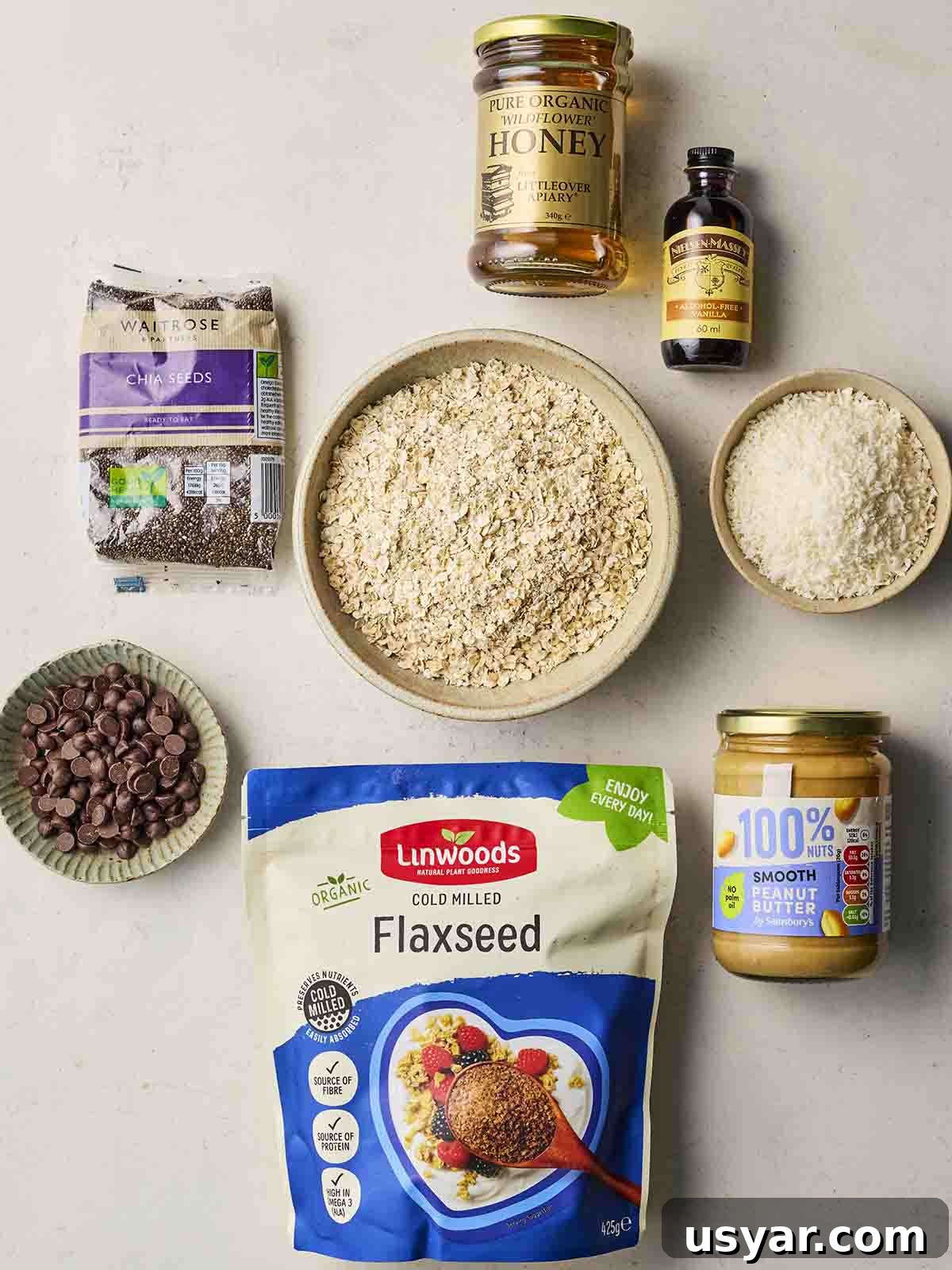 No-Bake Peanut Butter Oat Energy Balls Recipe 4 A collection of ingredients on a white surface, including oats, flaxseed, honey, dessicated coconut, peanut butter, chocolate chips, chia seeds and vanilla.