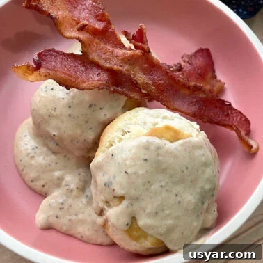 Classic Southern Sausage Gravy Recipe 14 Two biscuits with two strips of bacon topped with bacon gravy in a pink bowl.