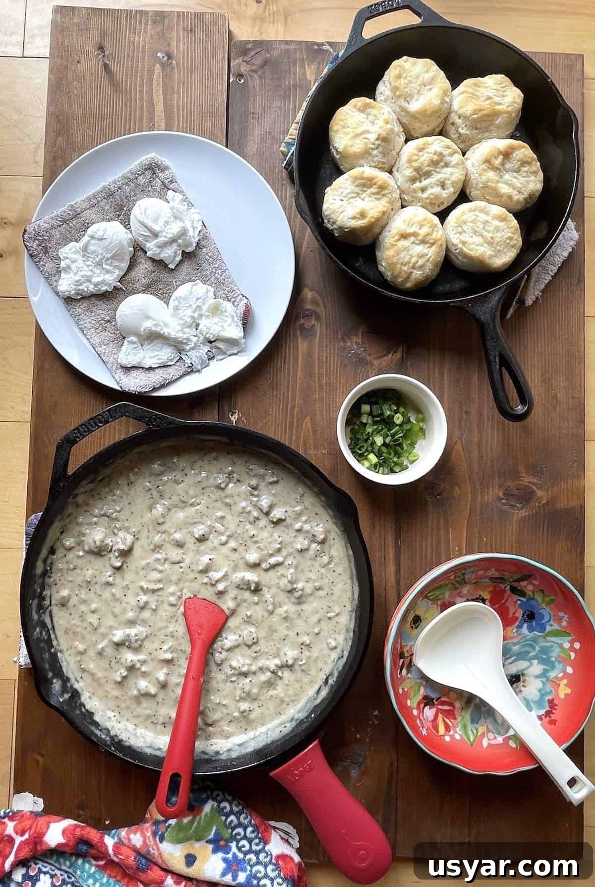 Classic Southern Sausage Gravy Recipe 4 A cast iron skillet filled with sausage gravy, a cast iron skillet of biscuits, poached eggs, and a bowl of green onions.