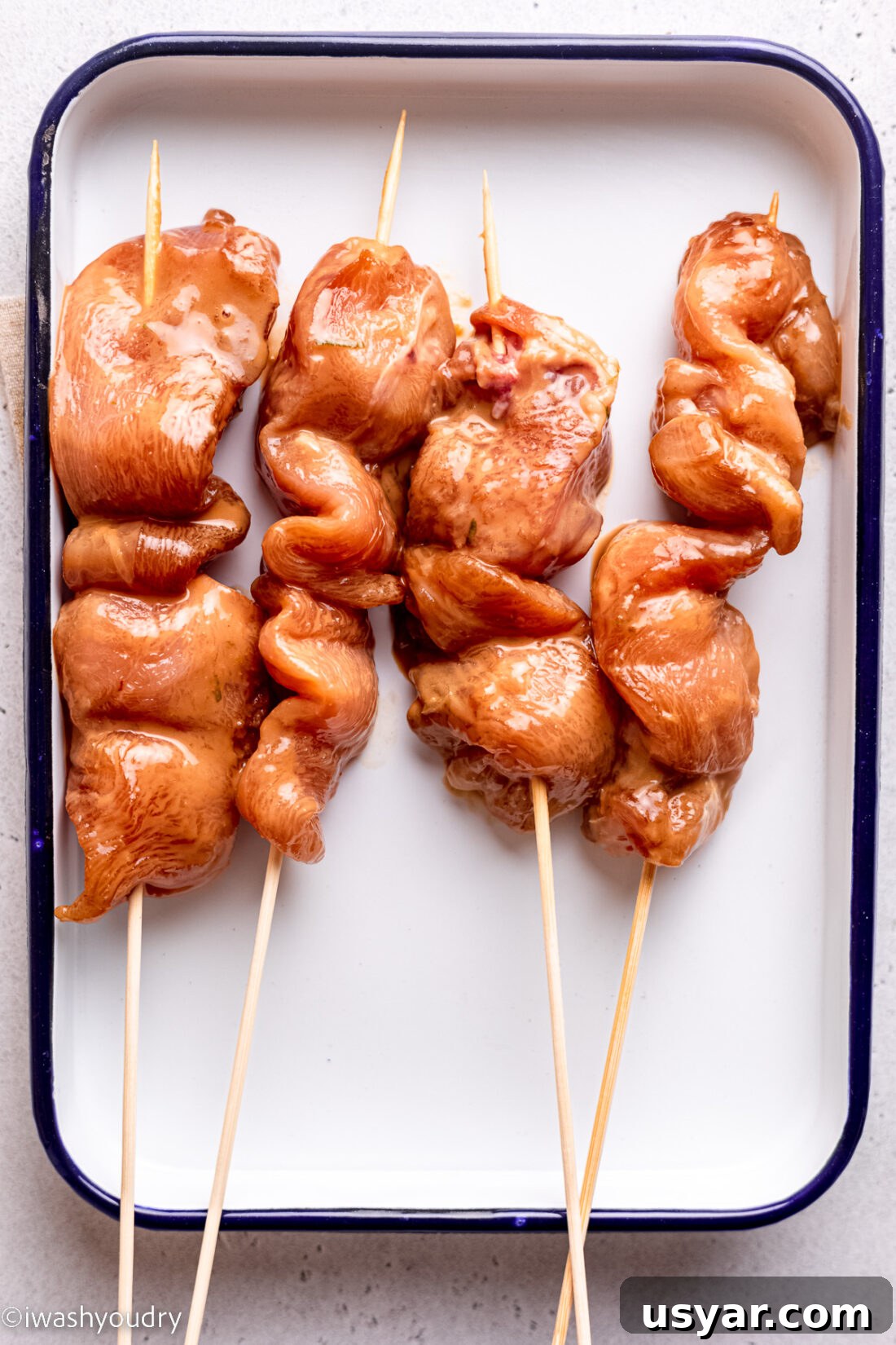 Thai Chicken Satay Recipe with Peanut Dipping Sauce 7 Raw chicken tenderloins on wooden skewers on white rectangle plate.
