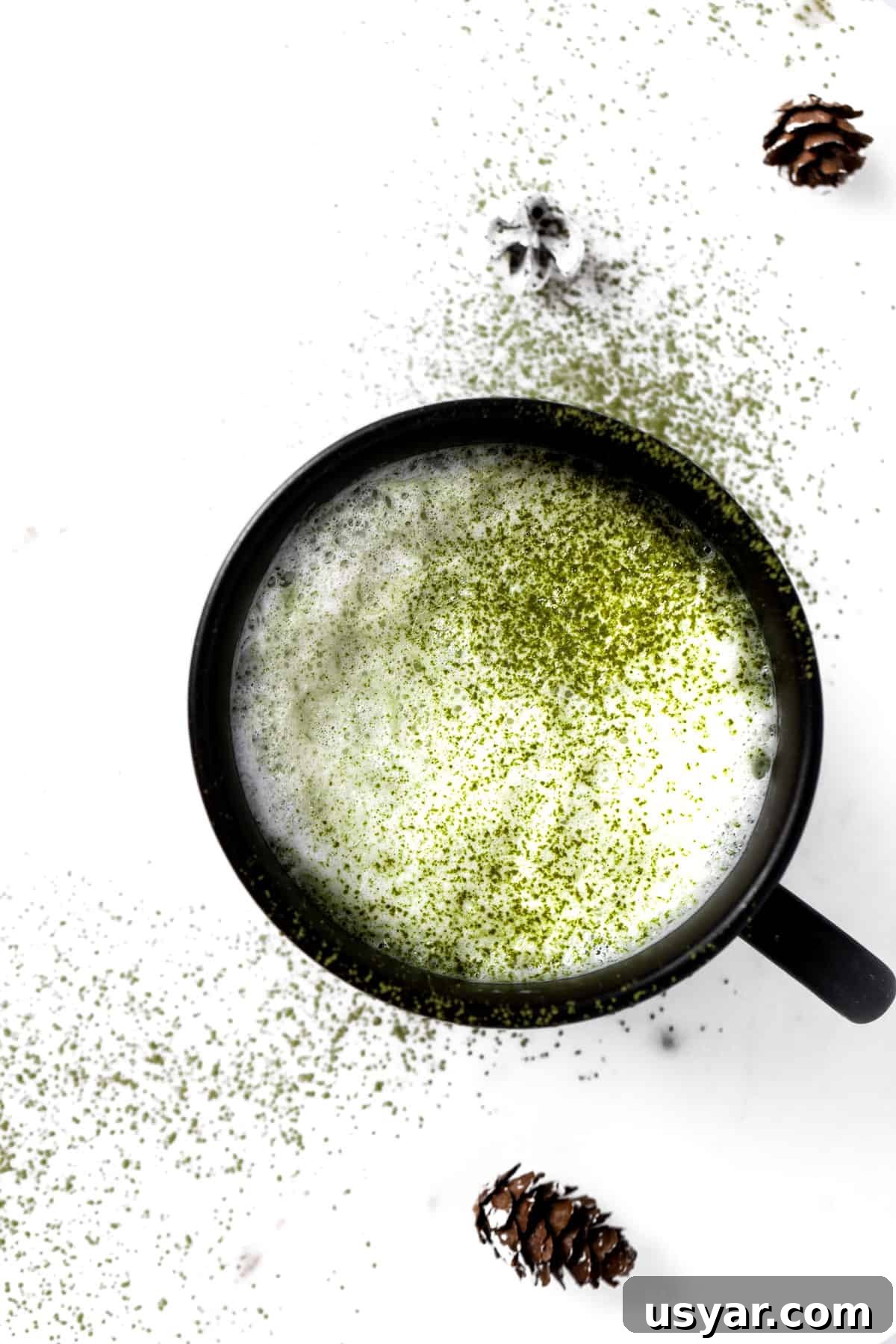Oat Milk Matcha Latte Recipe 7 A cup of oat milk matcha latte in a black mug, sprinkled with matcha powder on a white table.