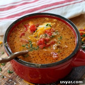 Beef Taco Soup with Ground Beef and Beans 6 Square side image of soup served in a red bowl on a wooden cutting board. Soup is garnished with sour cream, tomato, green onion, and cheddar cheese.
