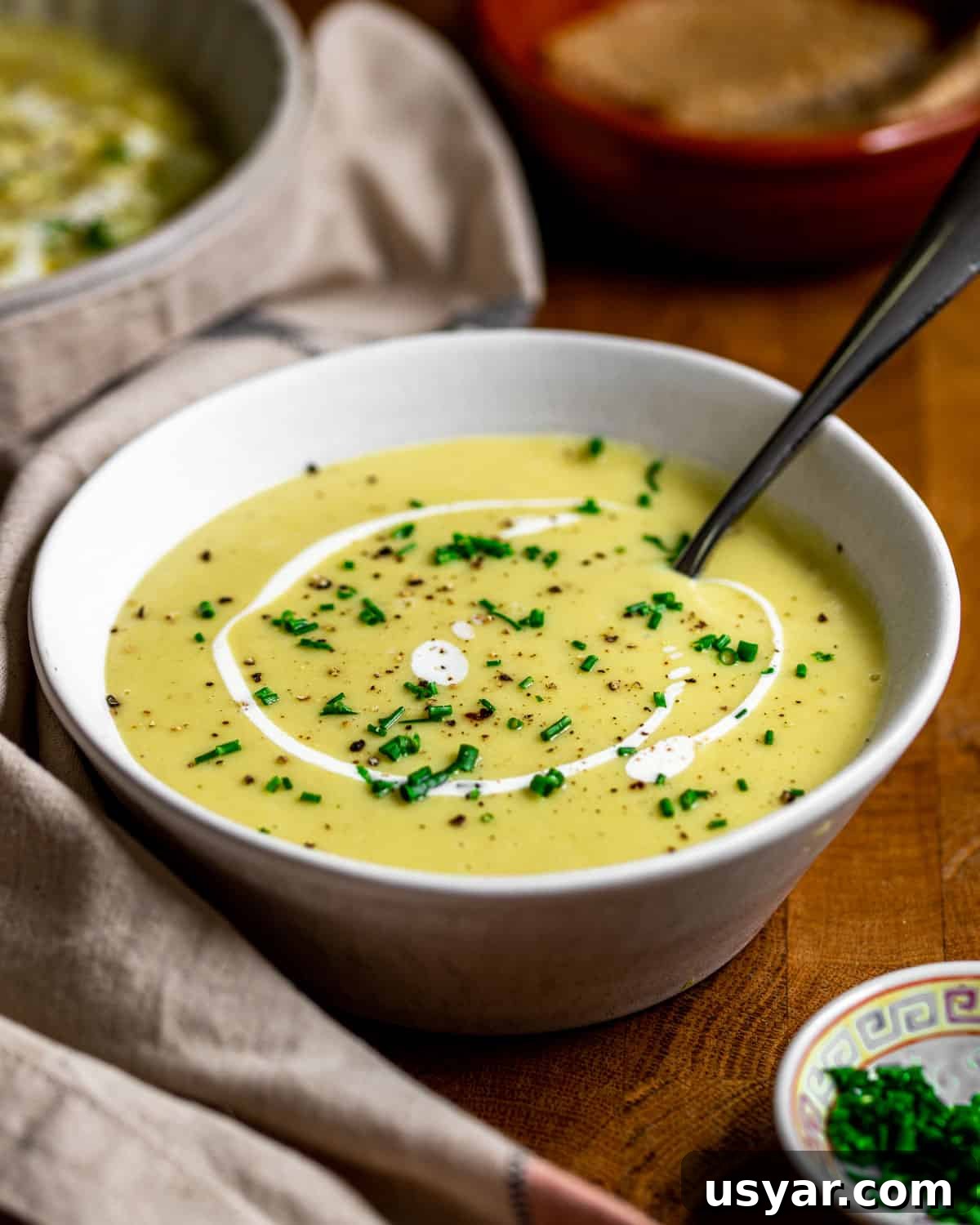 Creamy Vegan Leek and Potato Soup Vichyssoise 14 Photo of a smooth version of this vegan leek and potato soup recipe with a swirl of vegan cream and chopped fresh chives sprinkled on top, all on a wooden table with a napkin to the side.