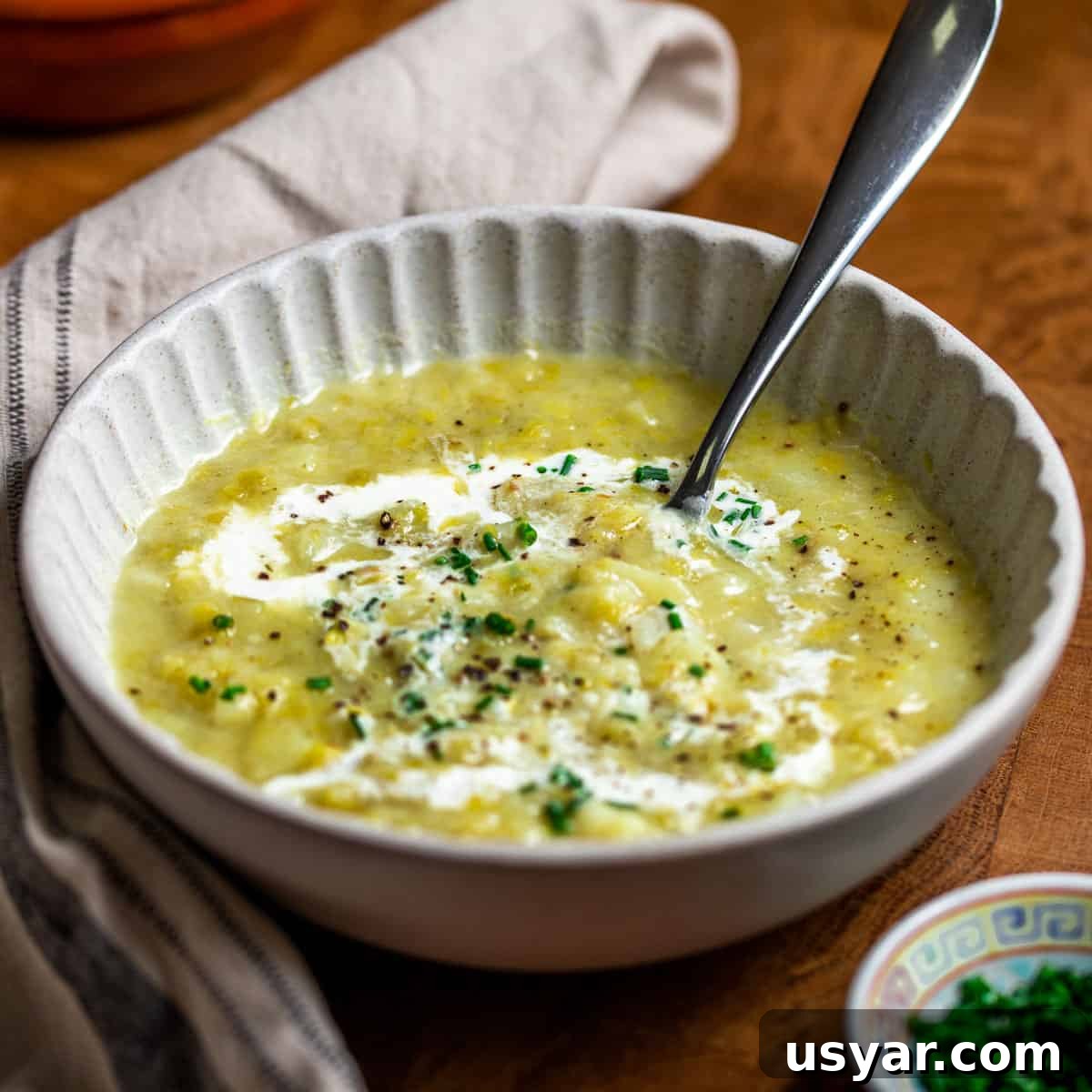 Creamy Vegan Leek and Potato Soup Vichyssoise 15 Photo of chunky vegan leek and potato soup in a cream bowl garnished with vegan cream and chives, with a napkin wrapped around the bowl, set on a wooden surface.