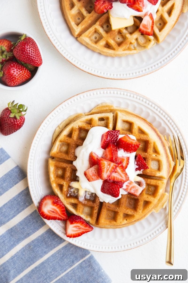 Top down photo of two plates of waffles with whipped cream, strawberries, and a blue striped napkin