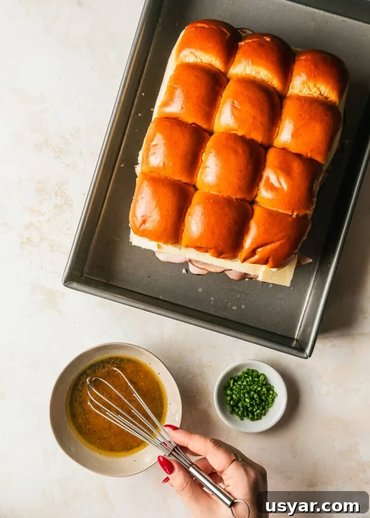 Oven Baked Brown Sugar Ham and Cheese Sliders 7 A hand whisking butter Dijon topping in a grey bowl next to a pan of sliders and a white bowl of chives on a tan counter.