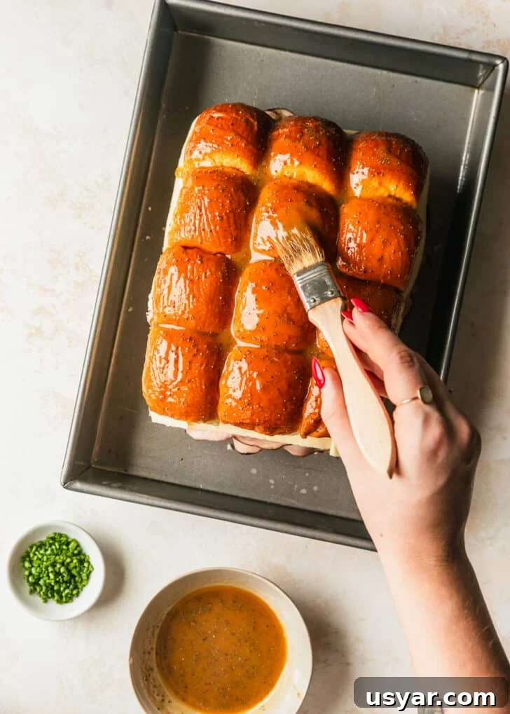 Oven Baked Brown Sugar Ham and Cheese Sliders 8 A hand using a pastry brush to brush brown sugar topping over ham and cheese sliders in a 9x13 pan on a tan counter.