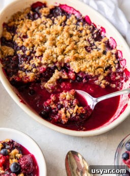 triple berry crisp in an oval baking dish with serving spoon holding some crisp