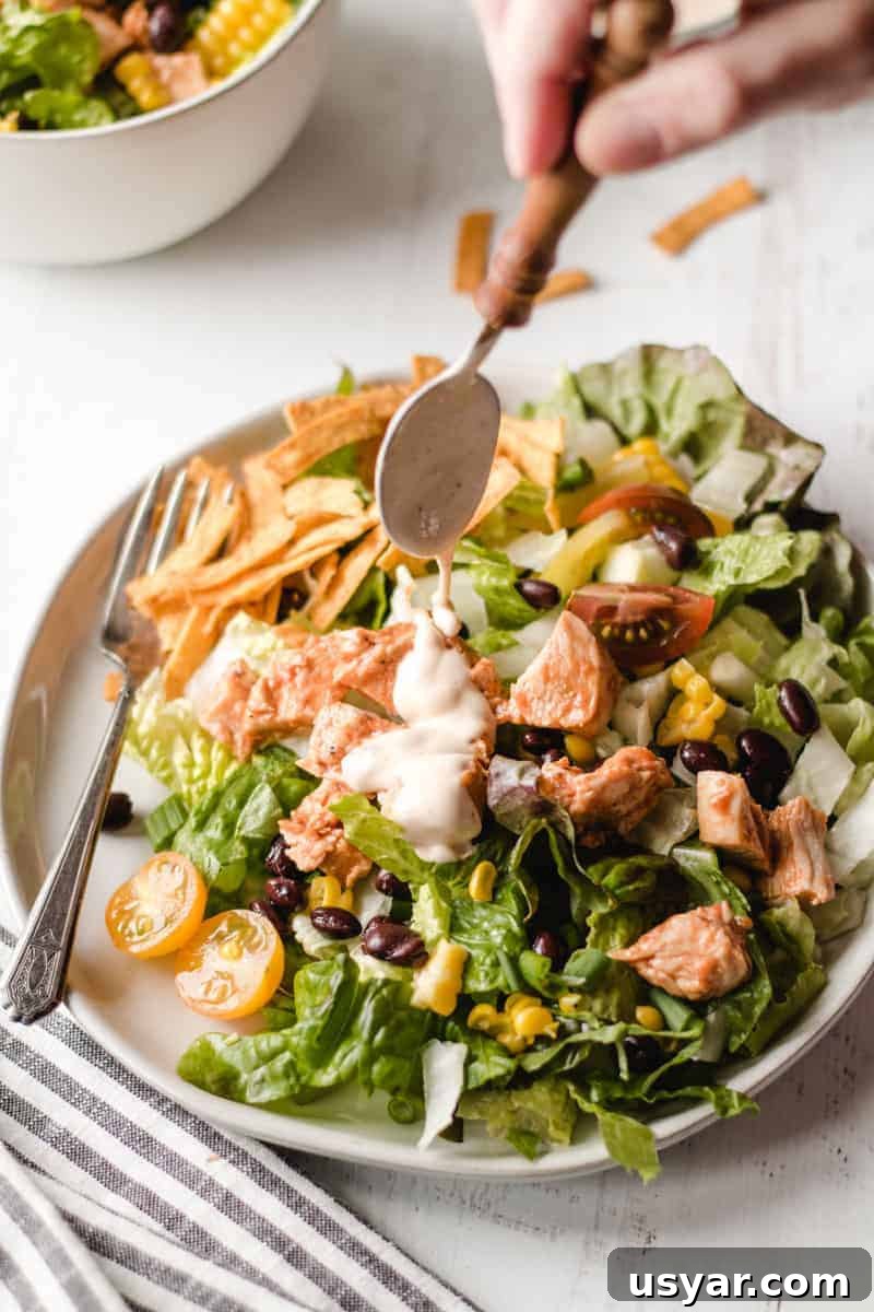 Pouring chipotle ranch dressing over a southwest bbq chicken salad piled high on a plate.