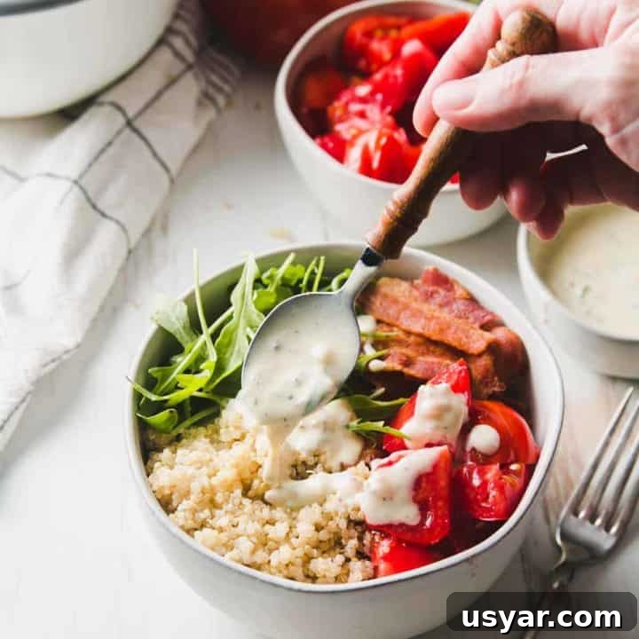 A bowl of quinoa, bacon, red tomatoes, and arugula is drizzled with yogurt dressing.