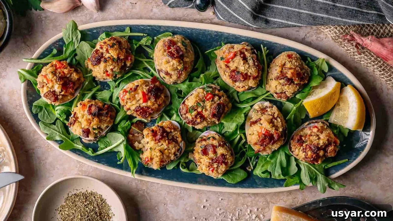 17 Potluck Appetizers to Get the Party Started 2 An oval platter with stuffed clams casino on a bed of fresh greens, garnished with lemon wedges, sits on a countertop with seasonings and kitchen linens nearby.