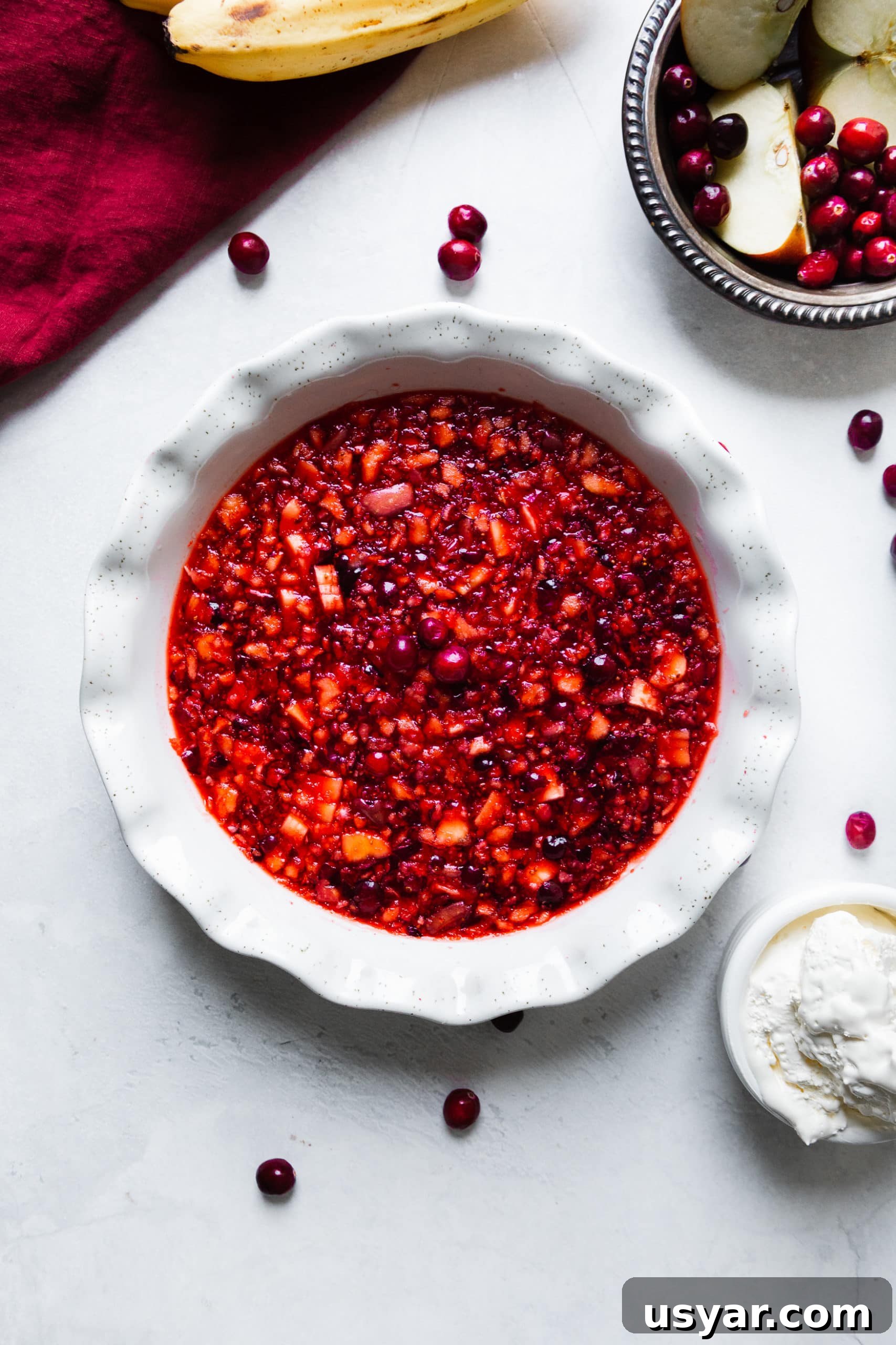 a photo taken directly above a white ceramic pie dish full of a chunky fruit jello. a small bowl of fresh whipped cream is sitting next to the dish.