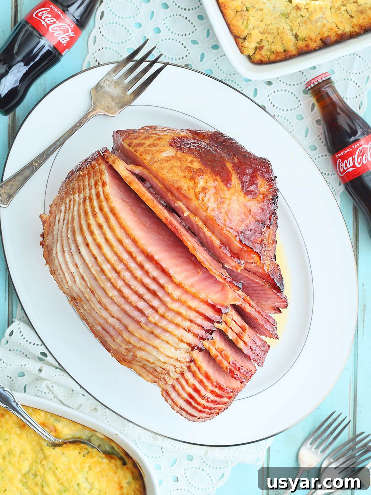 Coca Cola Glazed Ham Recipe 16 Sliced ham with Coca-Cola glaze on a white serving platter with side dishes and bottles of Coca Cola on the side.