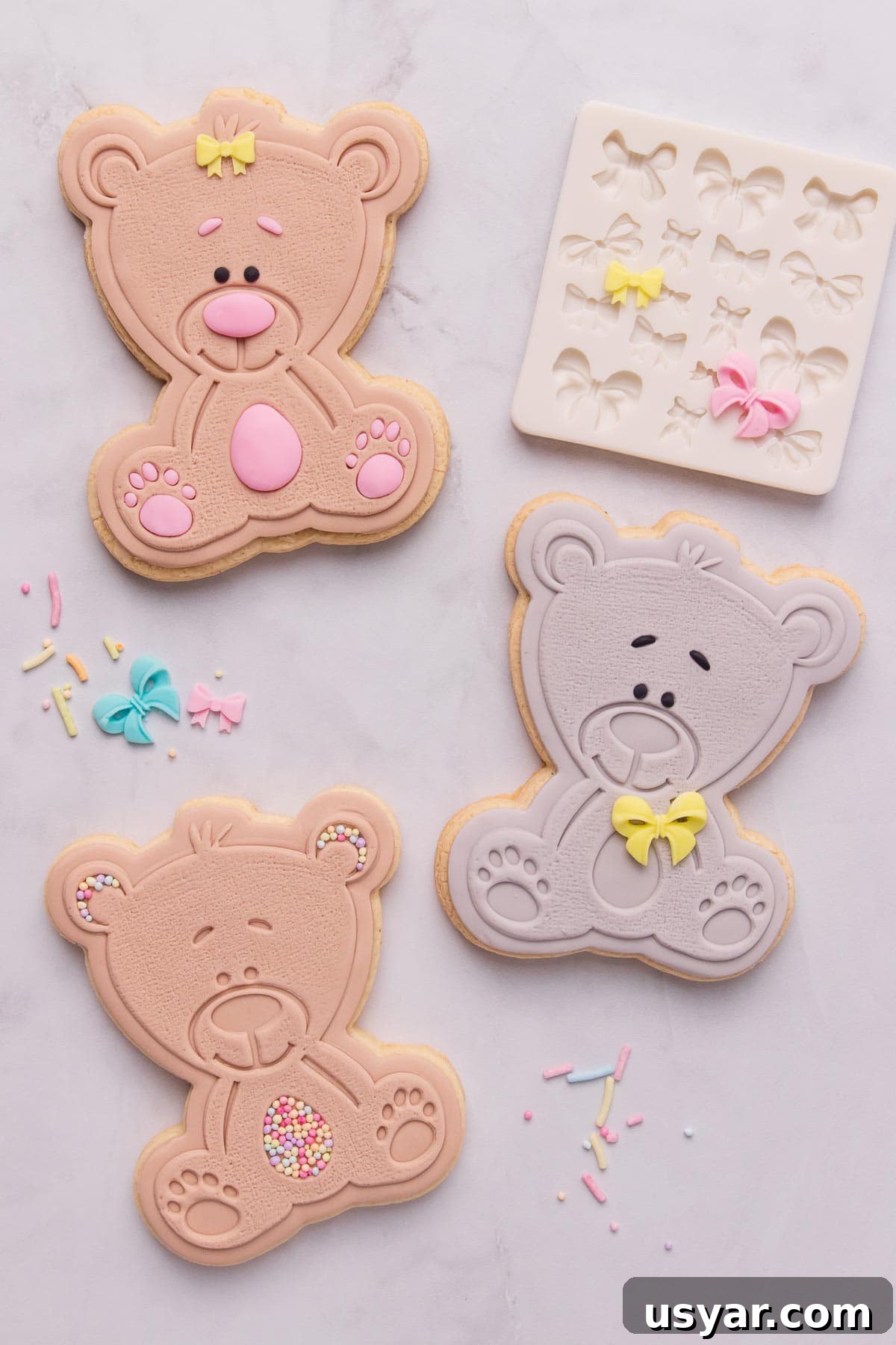 Three cute teddy bear shaped stamped fondant cookies, one bear is grey and the other two are light brown, and they're decorated with little bows and sprinkles.