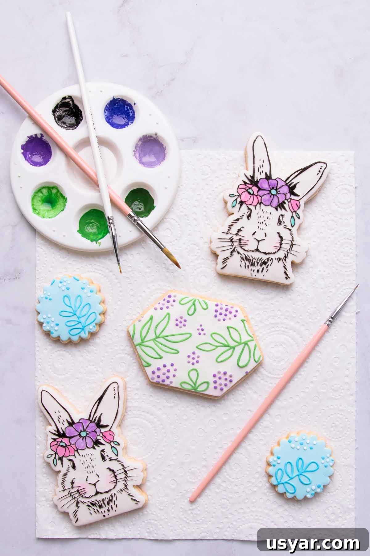 Two painted floral bunny cookies in pastel shades, with a leaf-patterned painted embossed cookie in shades of green, and two small blue cookies in the same leaf pattern, painted with blue and white paint.