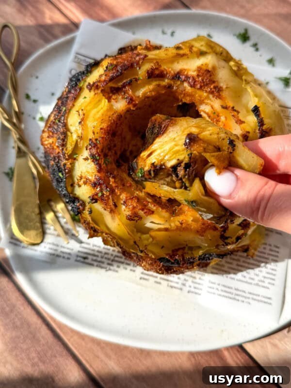 A hand pulls a crispy roasted slice from a whole roasted cabbage served on a white plate.