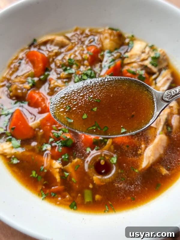 A close-up of No Water Chicken Soup with a spoonful of clear brown broth held above it, showing pieces of chicken, chopped carrots, and fresh herbs on top—perfect for a rich, waterless chicken soup recipe.