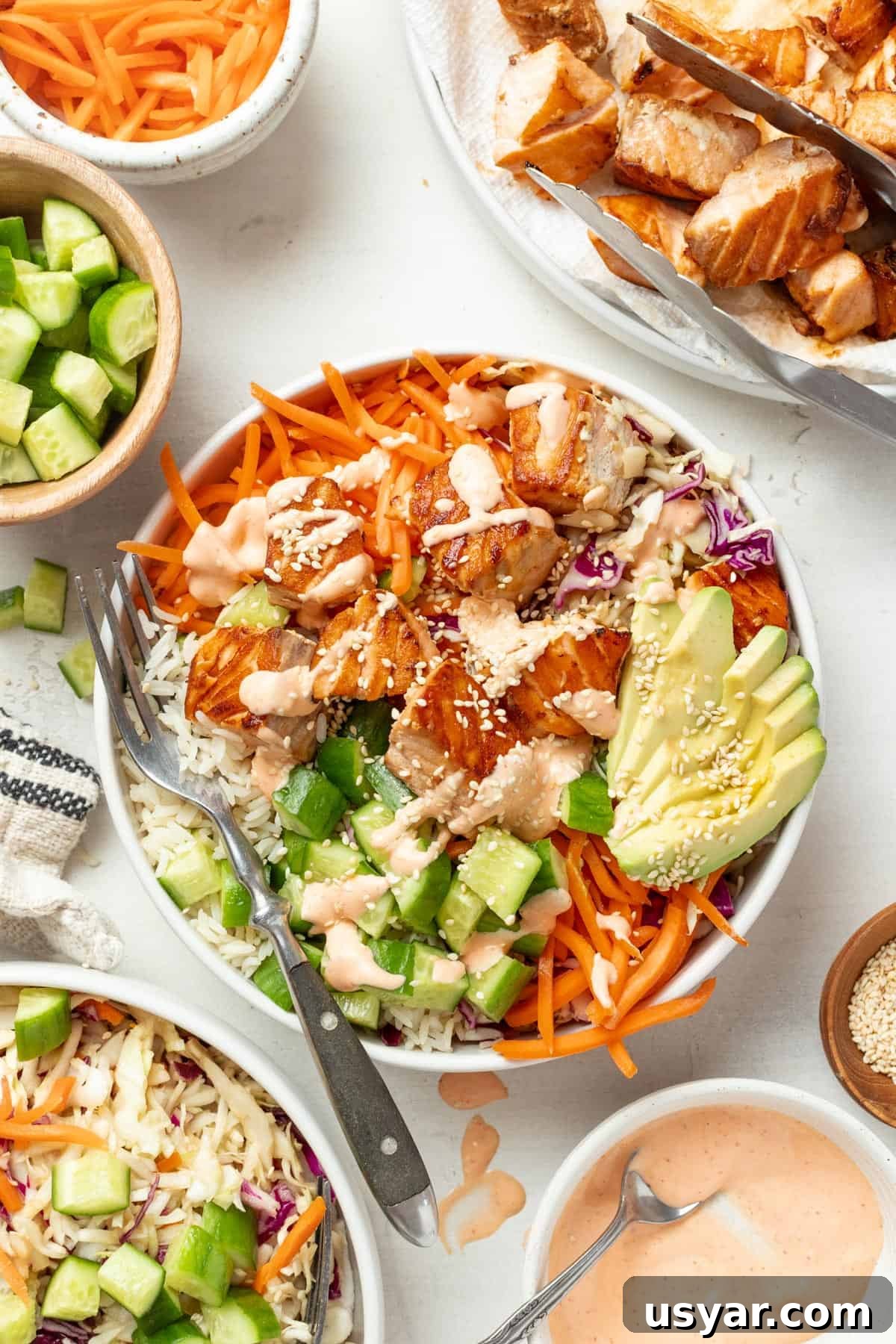 Spicy Mayo Salmon Rice Bowl 2 A salmon bowl with spicy mayo sauce, avocado, carrots, slaw, and rice on a table next to the ingredient components.