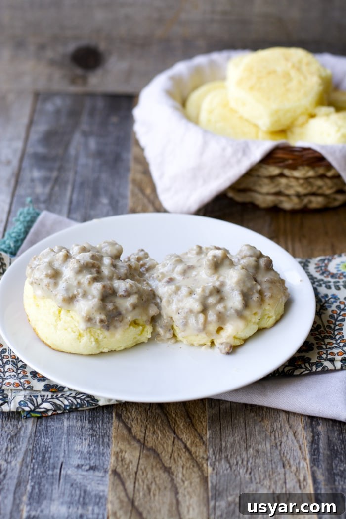 How to Make Gluten-Free Sausage Gravy 5 a biscuit split in half and topped with homemade sausage gravy on a white plate. A basket of biscuits rests in the background.