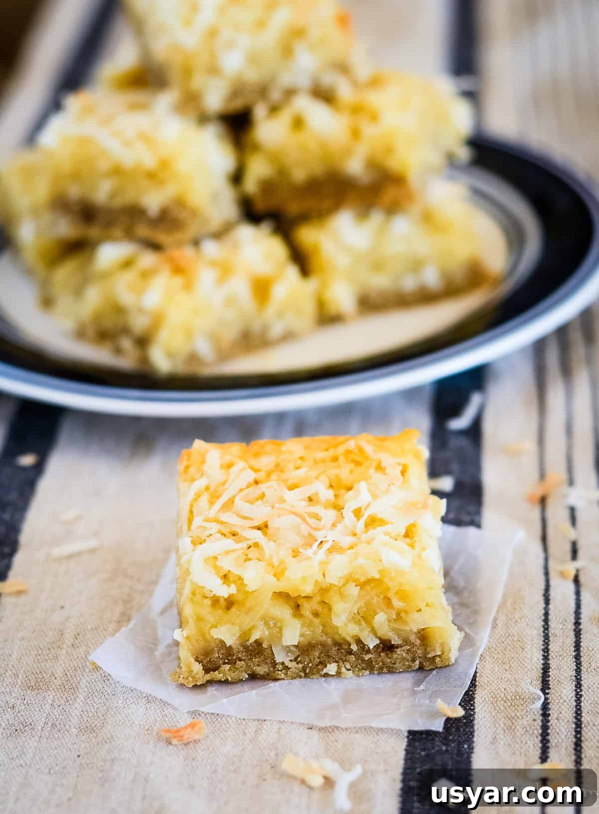 Homemade Chewy Buttery Coconut Bars 2 coconut bar on waxed paper with a plate of coconut bars in the background